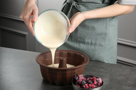 Woman pouring liquid dough from bowl into baking dish at gray table in kitchen, closeupの写真素材