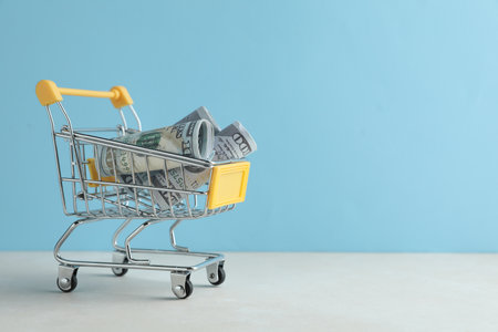 Saving money. Mini cart with dollar bills on white table against light blue background, space for textの写真素材