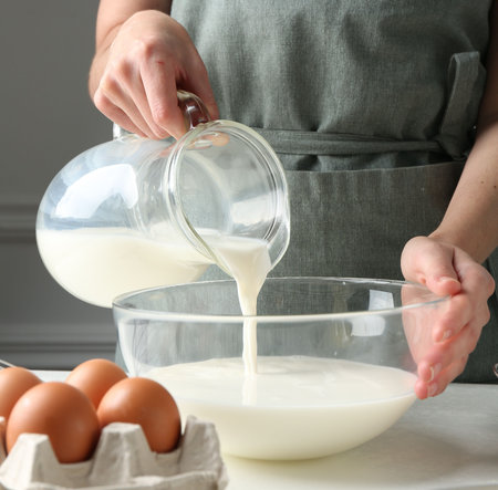 Making batter (liquid dough). Woman pouring milk into bowl at white table indoors, closeupの写真素材