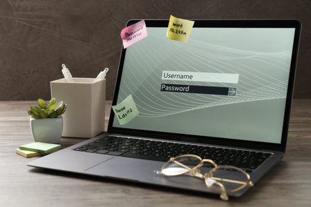 Laptop with password, username, paper notes, glasses and stationery on wooden table