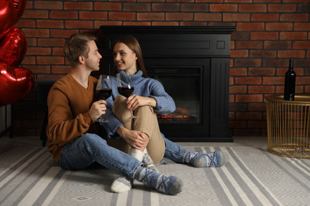 Lovely couple with glasses of wine near fireplace indoors, space for textの写真素材