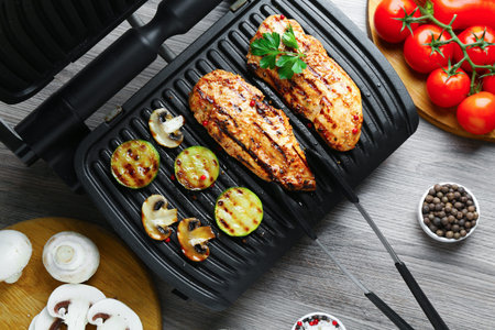 Electric grill with tasty chicken fillet, mushrooms, spices and vegetables on wooden table, flat layの写真素材