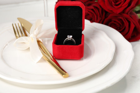 Romantic place setting. Box with engagement ring, plates, cutlery and roses on white table, closeup