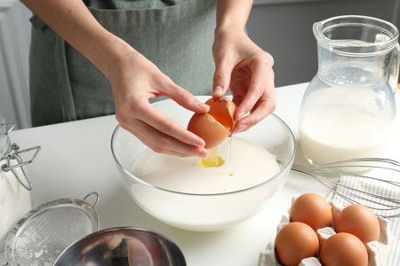 Making batter (liquid dough). Woman adding eggs into bowl at white table indoors, closeupの写真素材