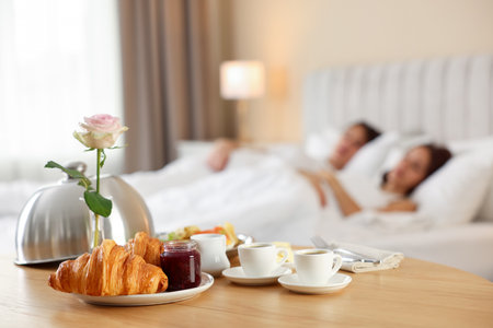 Delicious breakfast served on wooden table and couple sleeping in bed at hotel room, selective focusの写真素材