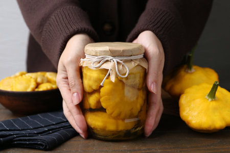 Woman with glass jar of pickled pattypan squashes, bowl and fresh ones on wooden table against light gray background, closeupの写真素材