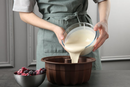 Woman pouring liquid dough from bowl into baking dish at gray table in kitchen, closeupの写真素材