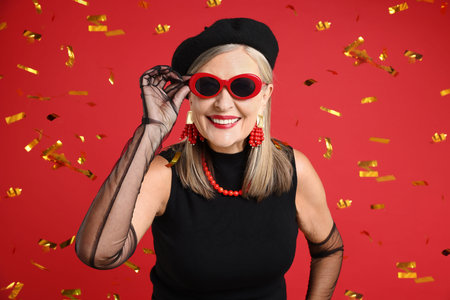 Happy senior woman in elegant outfit and flying confetti against red background