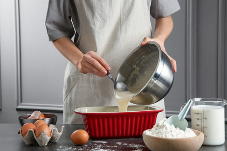 Woman pouring liquid dough from bowl into baking dish at gray table in kitchen, closeupの写真素材