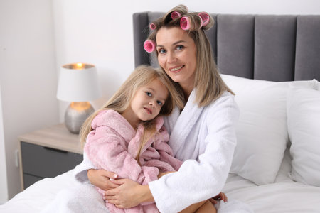 Mother and daughter in bathrobes with hair rollers on bed at homeの写真素材