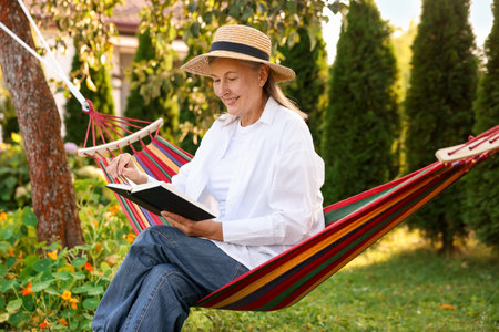 Senior woman with straw hat and book resting in hammock outdoorsの写真素材