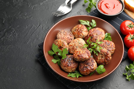 Delicious patties with parsley in bowl, tomatoes and ketchup on dark textured table, closeup. Space...
