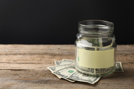 Jar with blank note and money on wooden table against black background. Space for textの写真素材