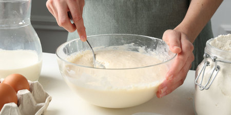 Woman making batter (liquid dough) at white table indoors, closeup