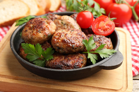Delicious patties with parsley and tomatoes in pan on wooden table, closeup