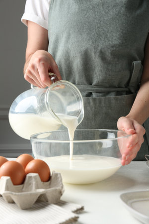 Making batter (liquid dough). Woman pouring milk into bowl at white table indoors, closeupの写真素材