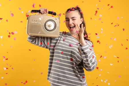 Excited young woman with radio receiver showing ok gesture among falling confetti on yellow backgroundの写真素材