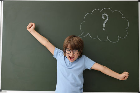 Back to school. Emotional boy near chalkboard with drawn thought bubble and question mark indoors