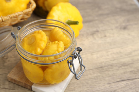 Pickled pattypan squashes in glass jar and fresh vegetables on wooden table, closeup. Space for textの写真素材