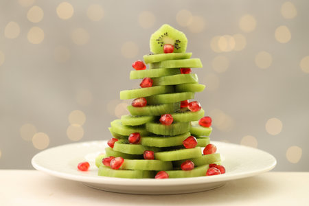 Christmas tree made of kiwi and pomegranate seeds on beige table against light background with blurred lights, closeup. Bokeh effectの写真素材