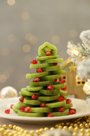 Christmas tree made of kiwi, pomegranate seeds and festive decor on table against light background with blurred lights, closeup. Bokeh effectの写真素材