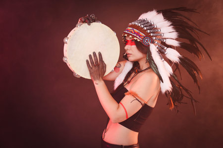 Shaman woman in native American Indian headdress with tambourine on dark background with smoke,...