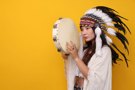 Shaman woman in native American Indian headdress with tambourine on orange background, space for...