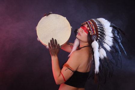Shaman woman in native American Indian headdress with tambourine on dark background with smokeの写真素材