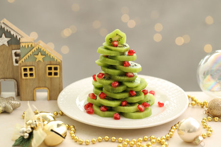 Christmas tree made of kiwi, pomegranate seeds and festive decor on beige table against light background with blurred lights, closeup. Bokeh effectの写真素材