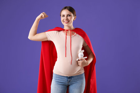 Pregnant woman with toy bunny wearing superhero cape on violet background