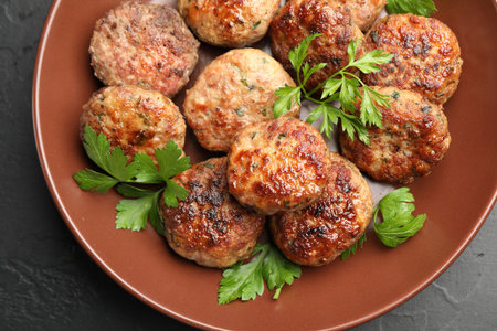Delicious patties with parsley in bowl on dark textured table, top view