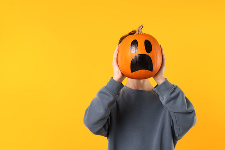 Halloween celebration. Boy holding pumpkin with drawn spooky face on orange background, space for textの写真素材
