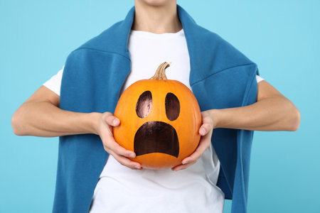 Halloween celebration. Boy holding pumpkin with drawn spooky face on light blue background, closeupの写真素材