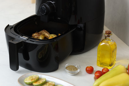 Air fryer basket with grilled chicken wings, potato wedges, vegetables and spices on white marble countertop, closeupの写真素材