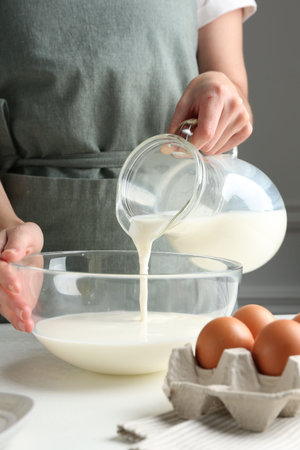 Making batter (liquid dough). Woman pouring milk into bowl at white table indoors, closeupの写真素材