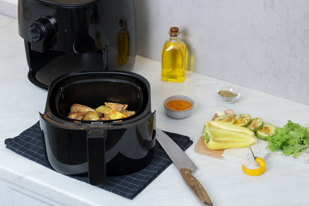Air fryer basket with grilled chicken wings, potato wedges, vegetables and spices on white marble countertop, closeupの写真素材