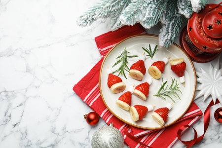 Christmas food. Santa hats made of marshmallows, strawberries, bananas, rosemary and decor on white marble table, flat lay. Space for textの写真素材