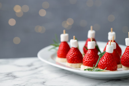 Christmas food. Santa hats made of marshmallows, strawberries, bananas and rosemary on white marble table against background with blurred lights, space for text. Bokeh effectの写真素材