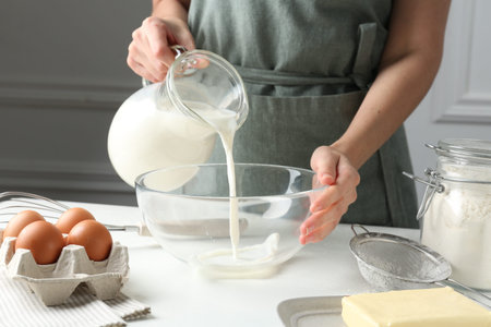 Making batter (liquid dough). Woman pouring milk into bowl at white table indoors, closeup