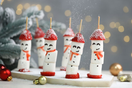 Sprinkling powdered sugar onto funny snowmen made of marshmallows, strawberries and Christmas decor at table against background with blurred lights, closeup. Bokeh effectの写真素材