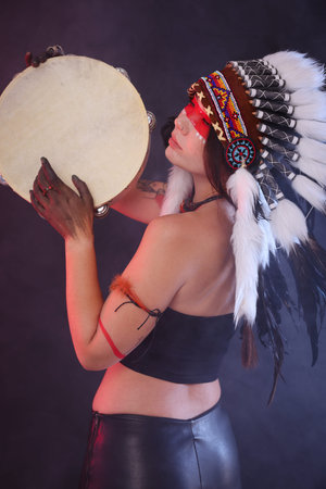 Shaman woman in native American Indian headdress with tambourine on dark background with smokeの写真素材