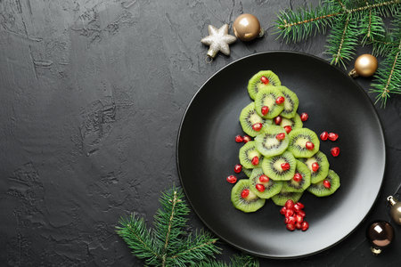 Christmas tree made of kiwi, pomegranate seeds and festive decor on black table, flat lay. Space for textの写真素材