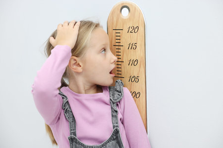 Surprised little girl measuring her height with chart near light grey wall