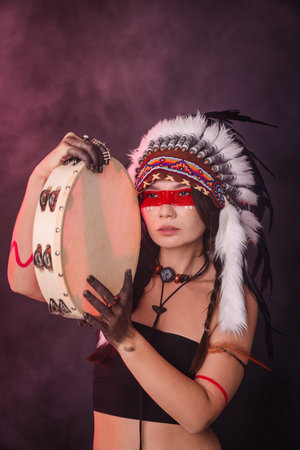 Shaman woman in native American Indian headdress with tambourine on dark background with smoke