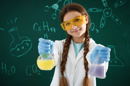 Back to school. Girl with flasks of liquids against chalkboard with chemical formulas indoorsの写真素材