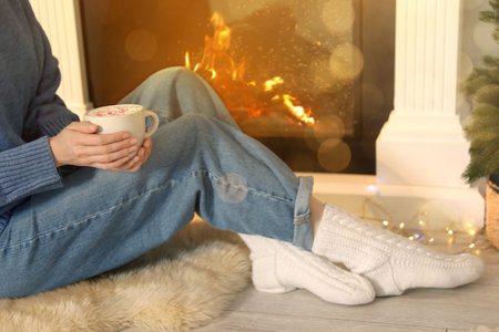Woman with tasty hot cocoa by fireplace at home, closeup. Christmas vibes