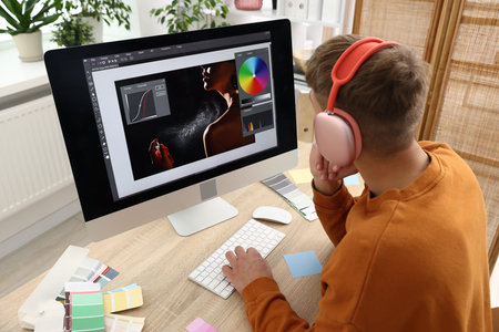 Professional retoucher with headphones editing photo on computer at table in office, back view