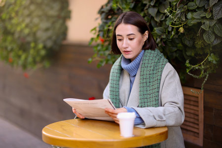 Beautiful woman in coat and scarf reading newspaper at table in outdoor cafeの写真素材