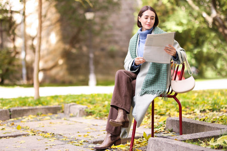 Beautiful woman in coat and scarf with bag reading newspaper on chair in autumn park. Space for textの写真素材