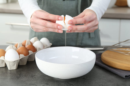 Woman cracking egg into bowl at gray table in kitchen, closeup